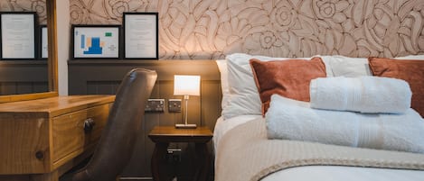 Habitación de hotel con cama, escritorio, silla, mesita de noche con lámpara y un panel con diplomas enmarcados.