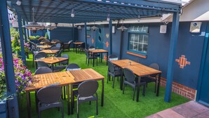 Un espace extérieur aménagé en terrasse avec des tables et des chaises, une pergola agrémentée de guirlandes lumineuses et un mur de briques avec une fenêtre.