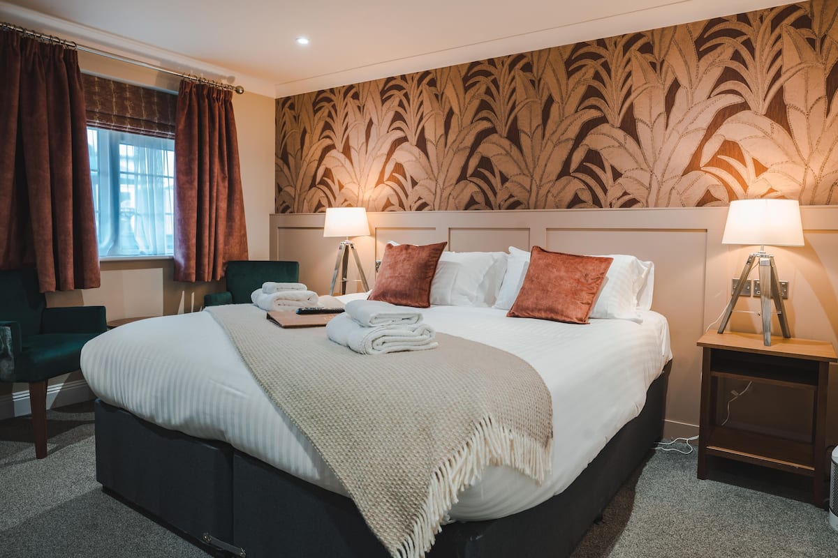 A hotel room with a large bed, two bedside lamps, a green armchair, a small table with a towel and remote, and a patterned wallpaper.