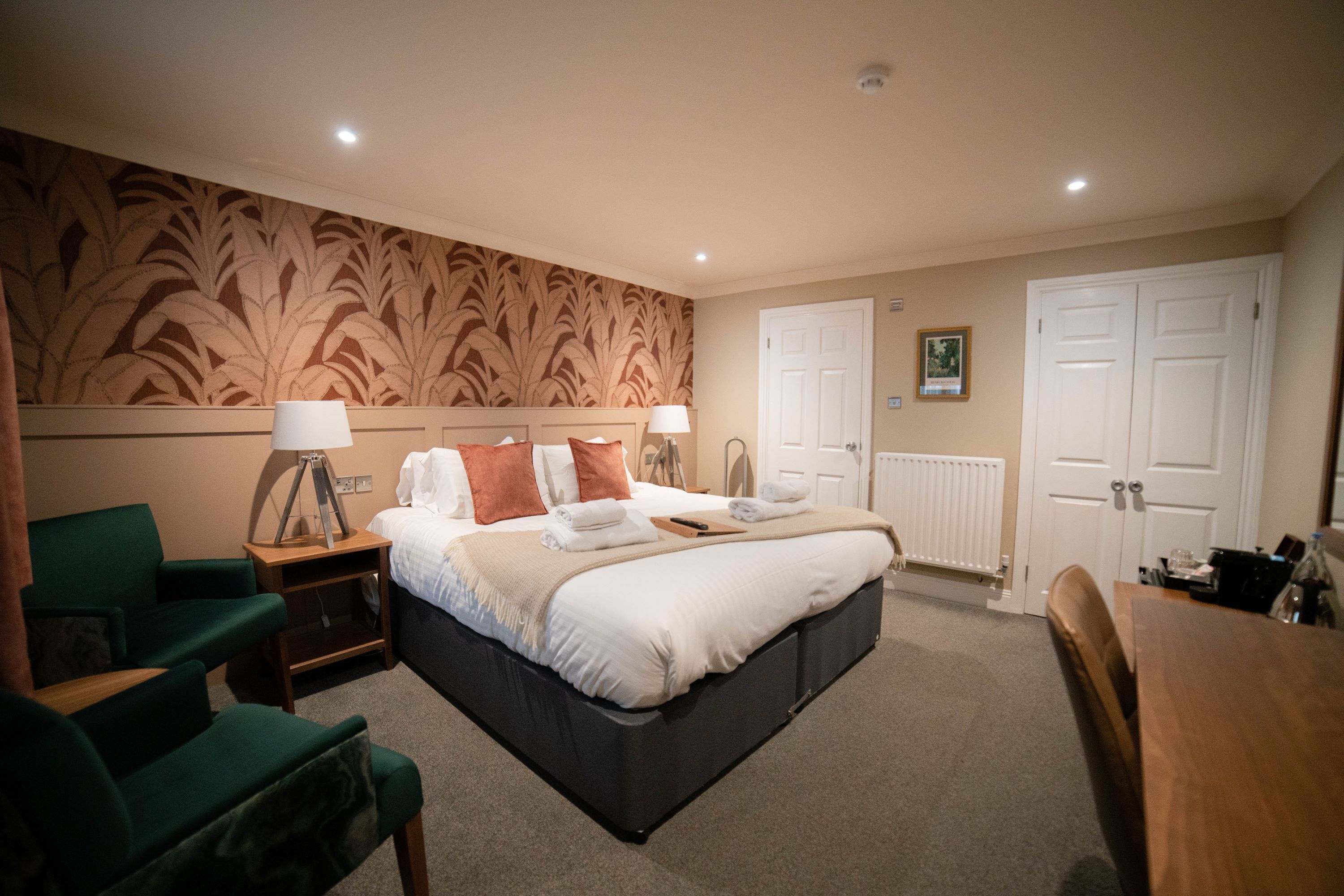 A hotel room with a large bed, two bedside tables with lamps, a green armchair, a wooden desk, and a radiator.