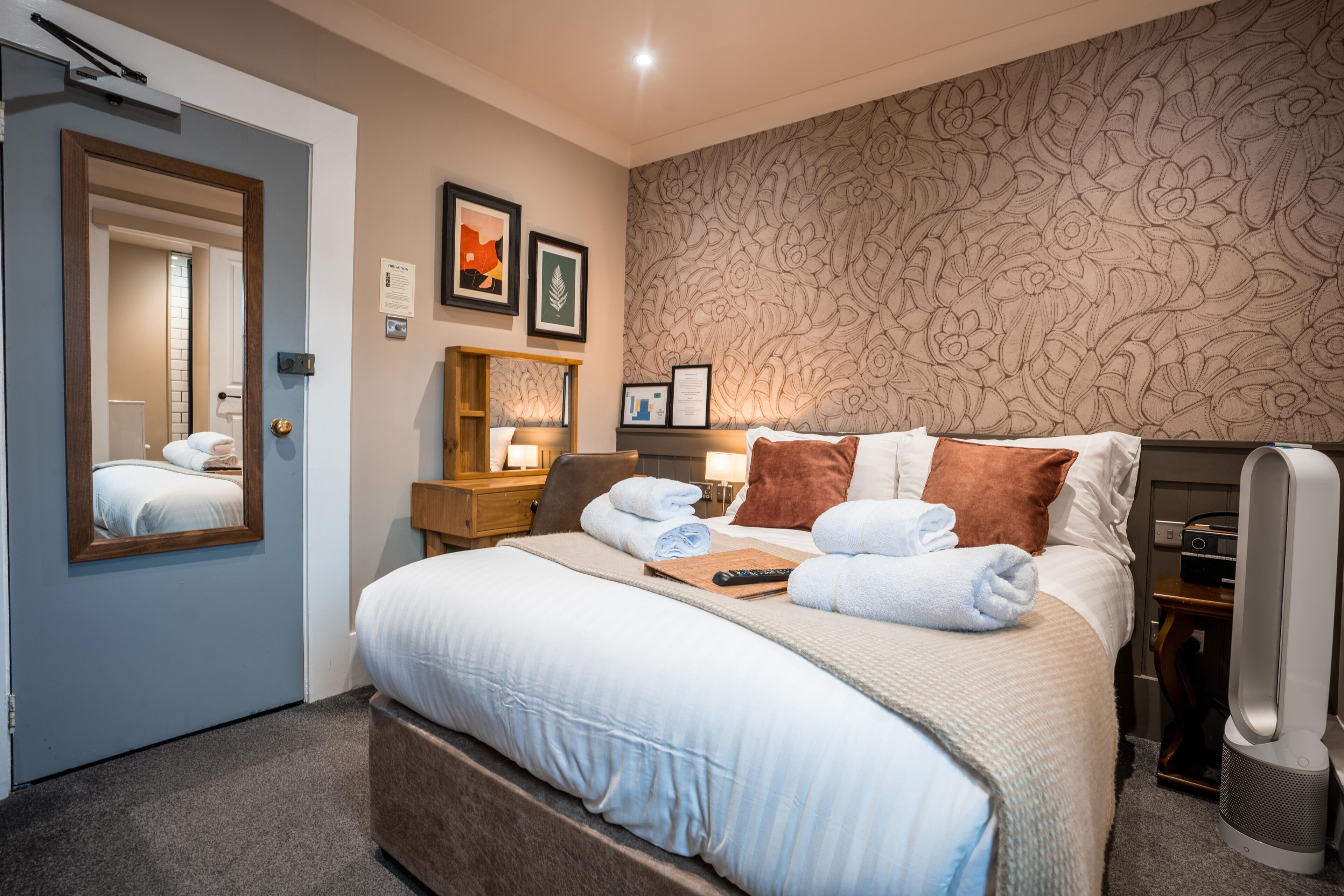 A hotel room with a large bed, a bedside table with a lamp, a mirror, and a floral-patterned wall.