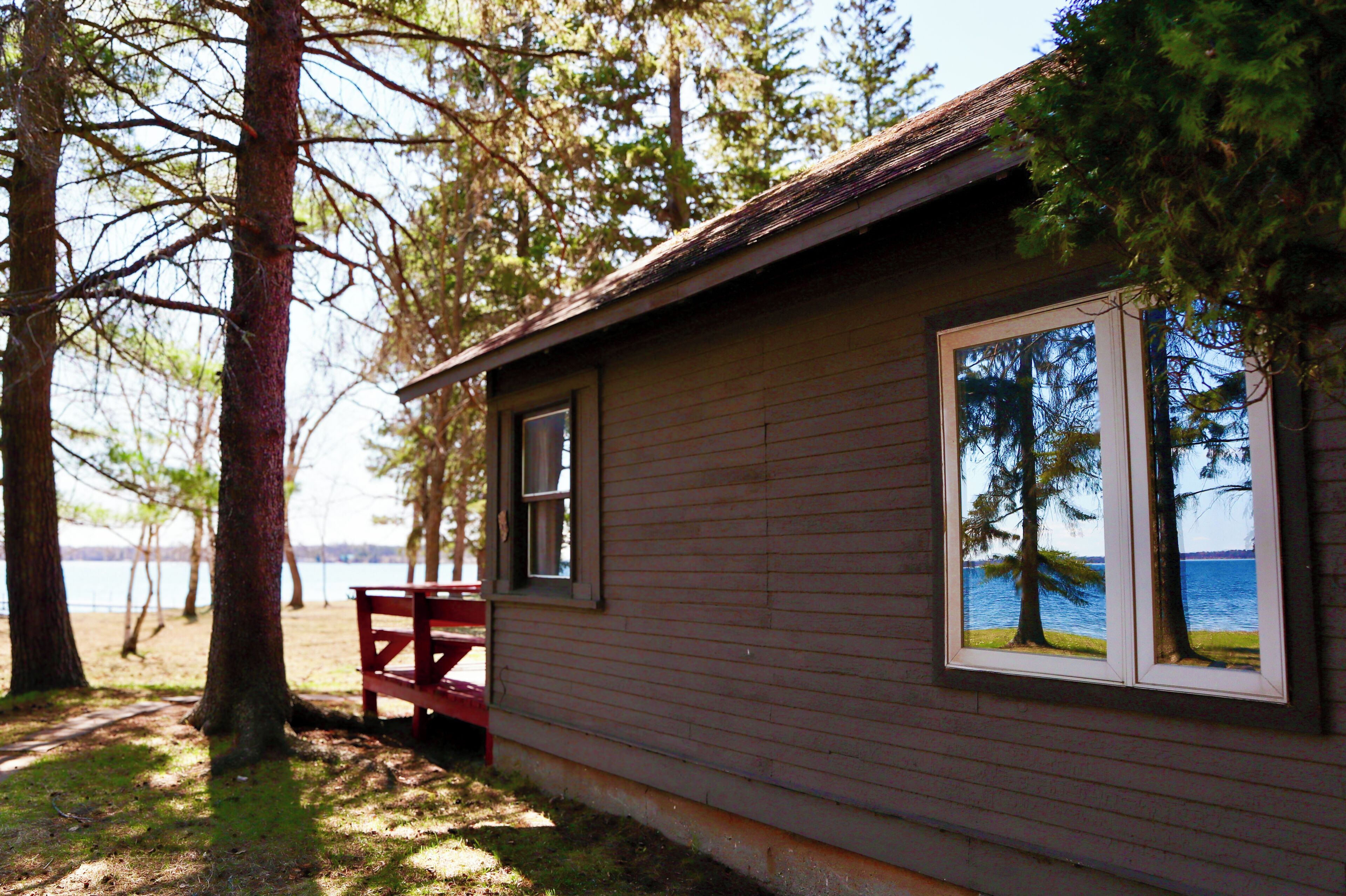A cabin with a view of the lake, a bench, and a window reflecting trees.