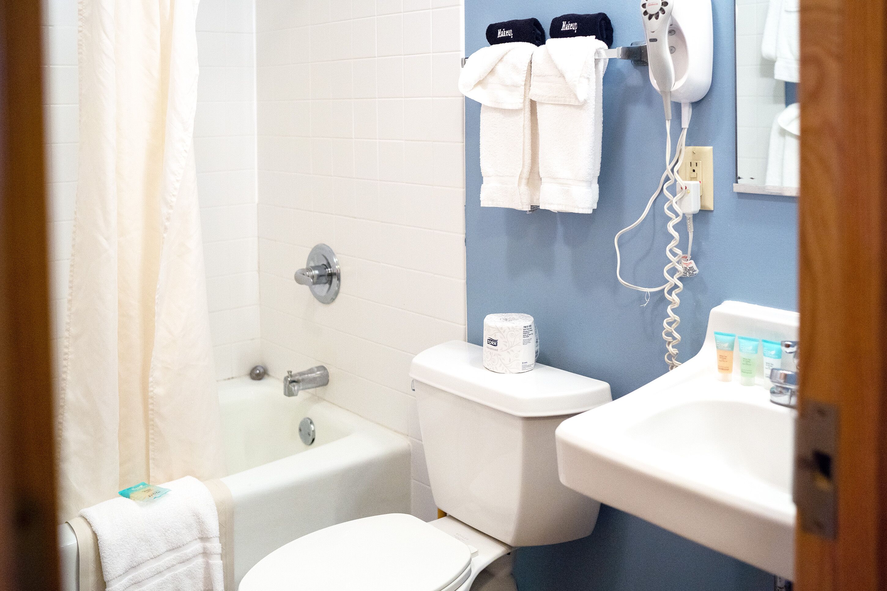 Un baño con bañera, inodoro y lavabo. Hay toallas colgadas en ganchos y un secador de pelo en la pared.