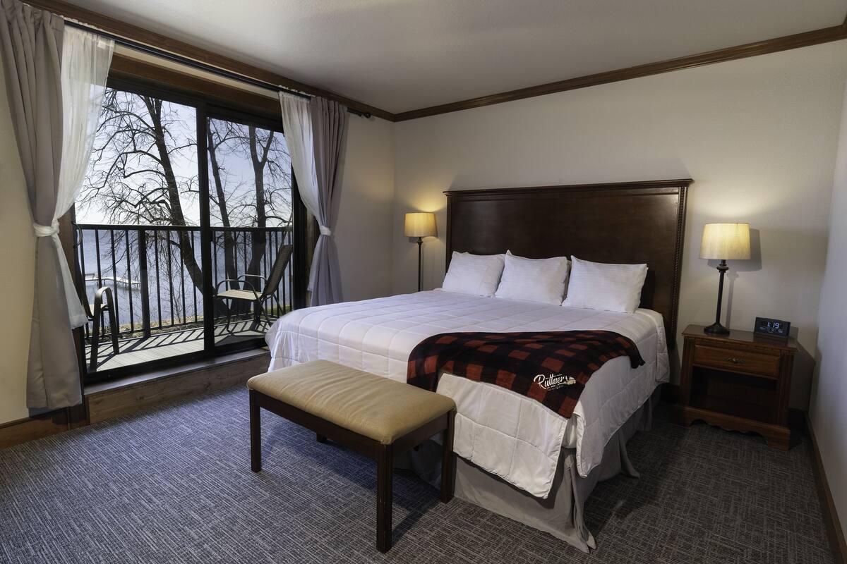 Hotelkamer met een groot bed, twee bedlampen, een bankje, een nachtkastje en uitzicht op bomen door een balkongevel.