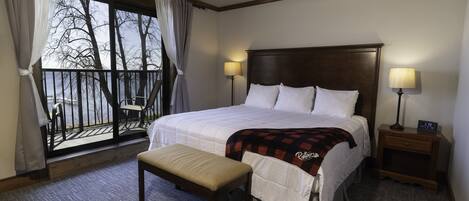 A hotel room with a large bed, two bedside lamps, a bench, a nightstand, and a view of trees through a balcony window.