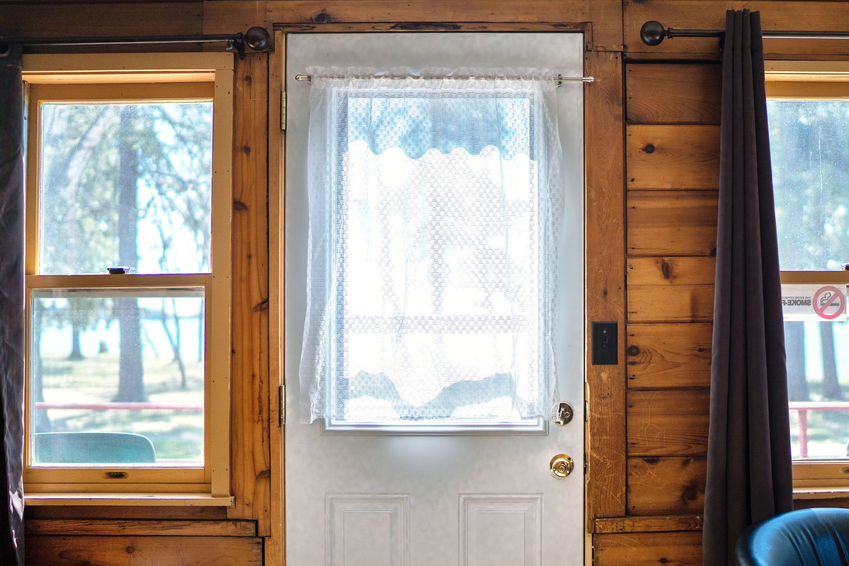 Una puerta de madera con un timbre blanco, una ventana con cortina de encaje y vistas a árboles fuera.