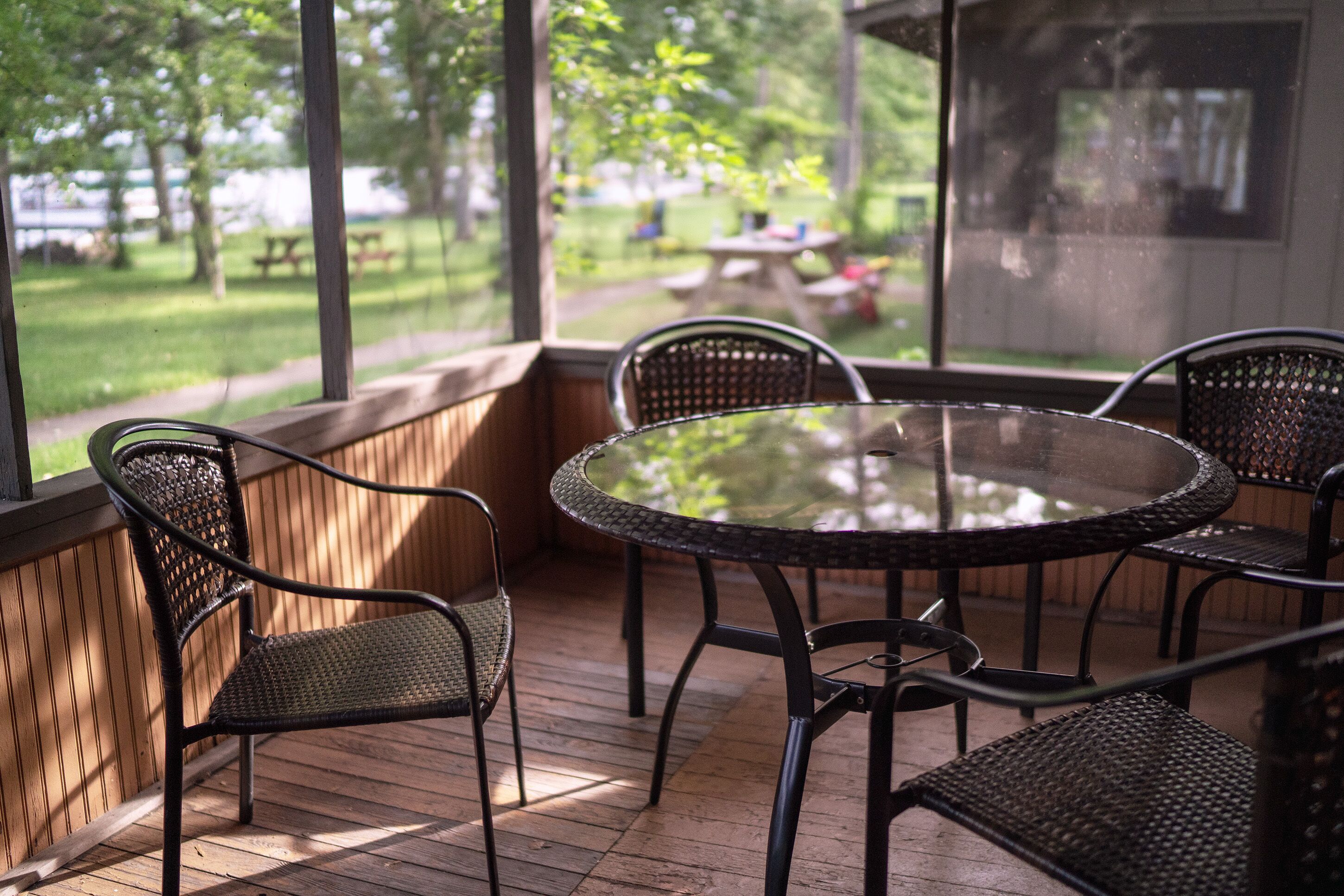 A covered patio with a round glass table and four wicker chairs, overlooking a grassy area with picnic tables.