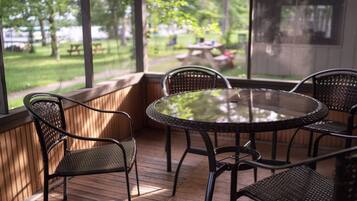 A covered patio with a round glass table and four wicker chairs, overlooking a grassy area with picnic tables.
