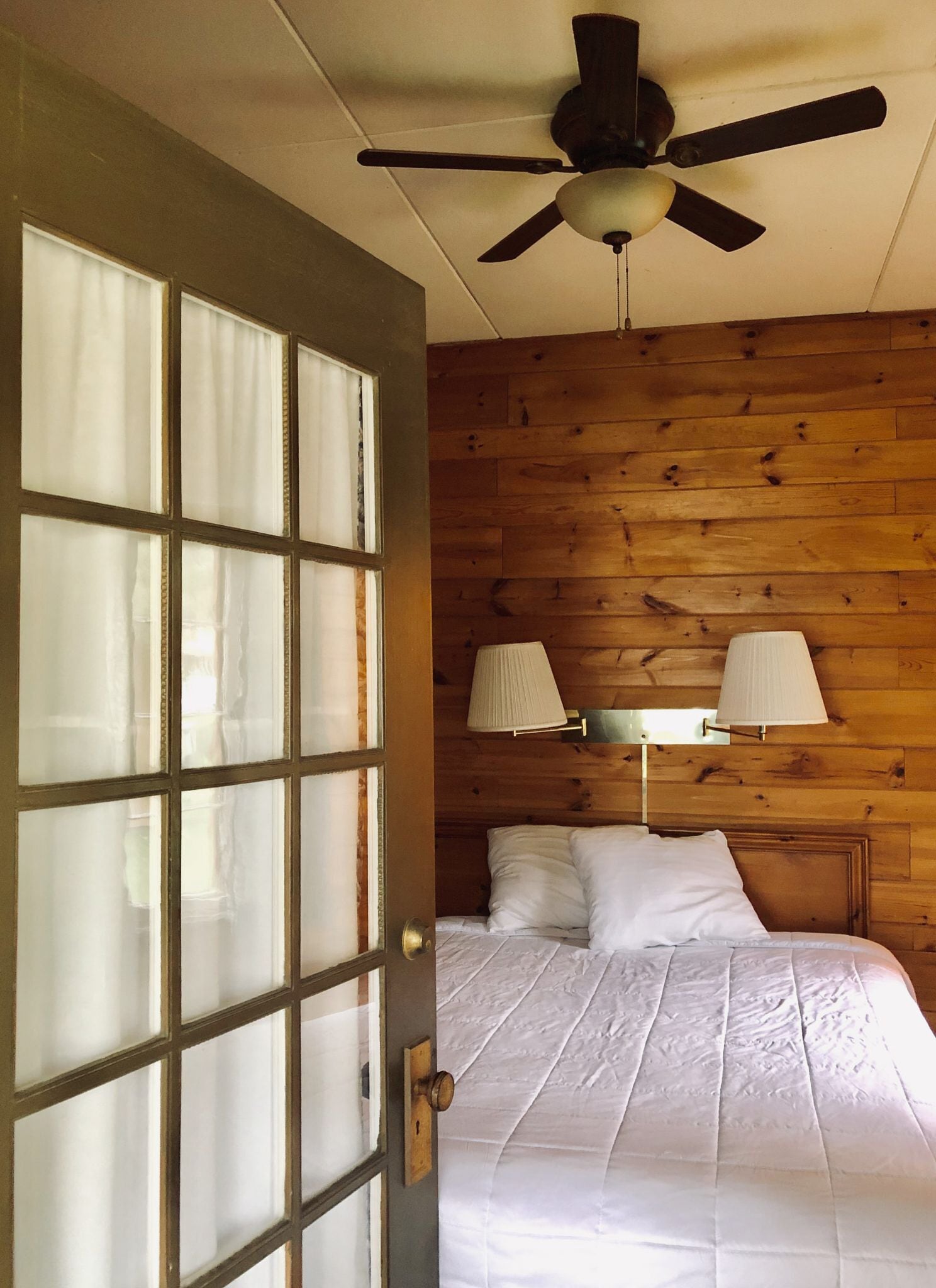 A bedroom with a wooden wall, a bed with white bedding, a ceiling fan, and two wall-mounted lamps.