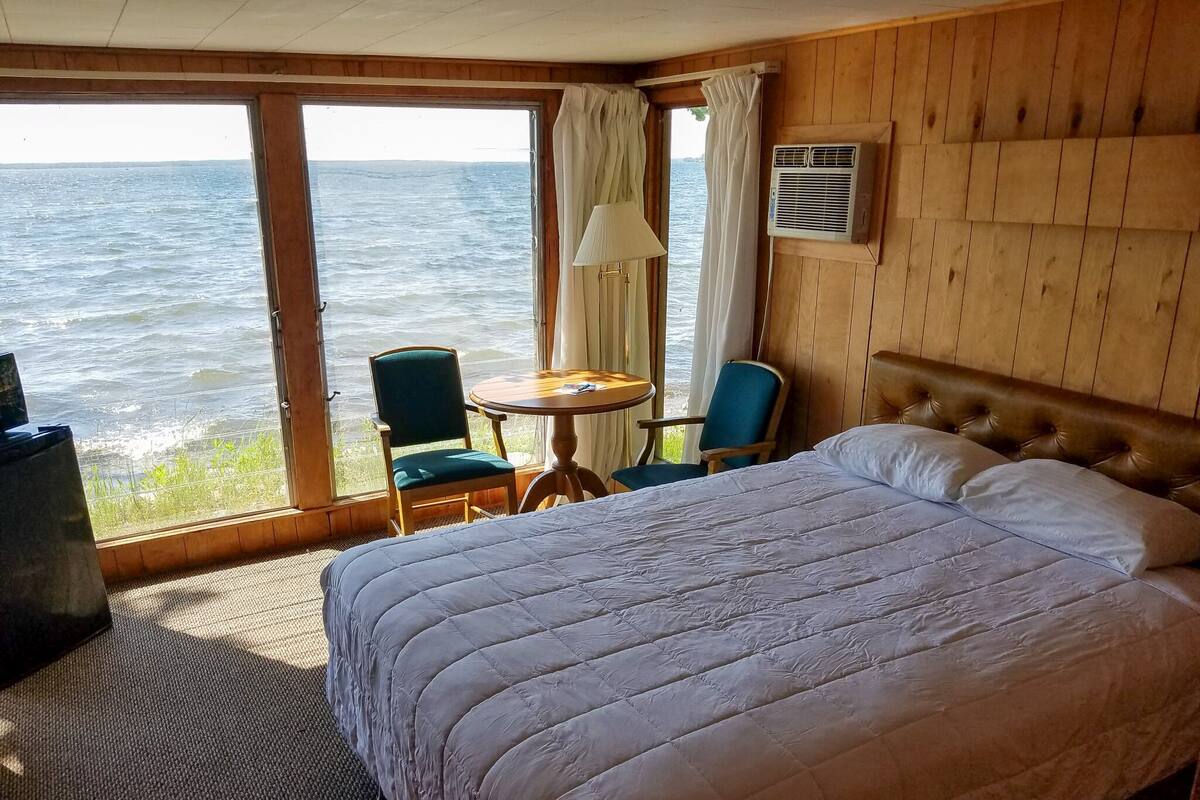 Een kamer met een groot bed, twee stoelen, een tafeltje en uitzicht op zee door een groot raam.