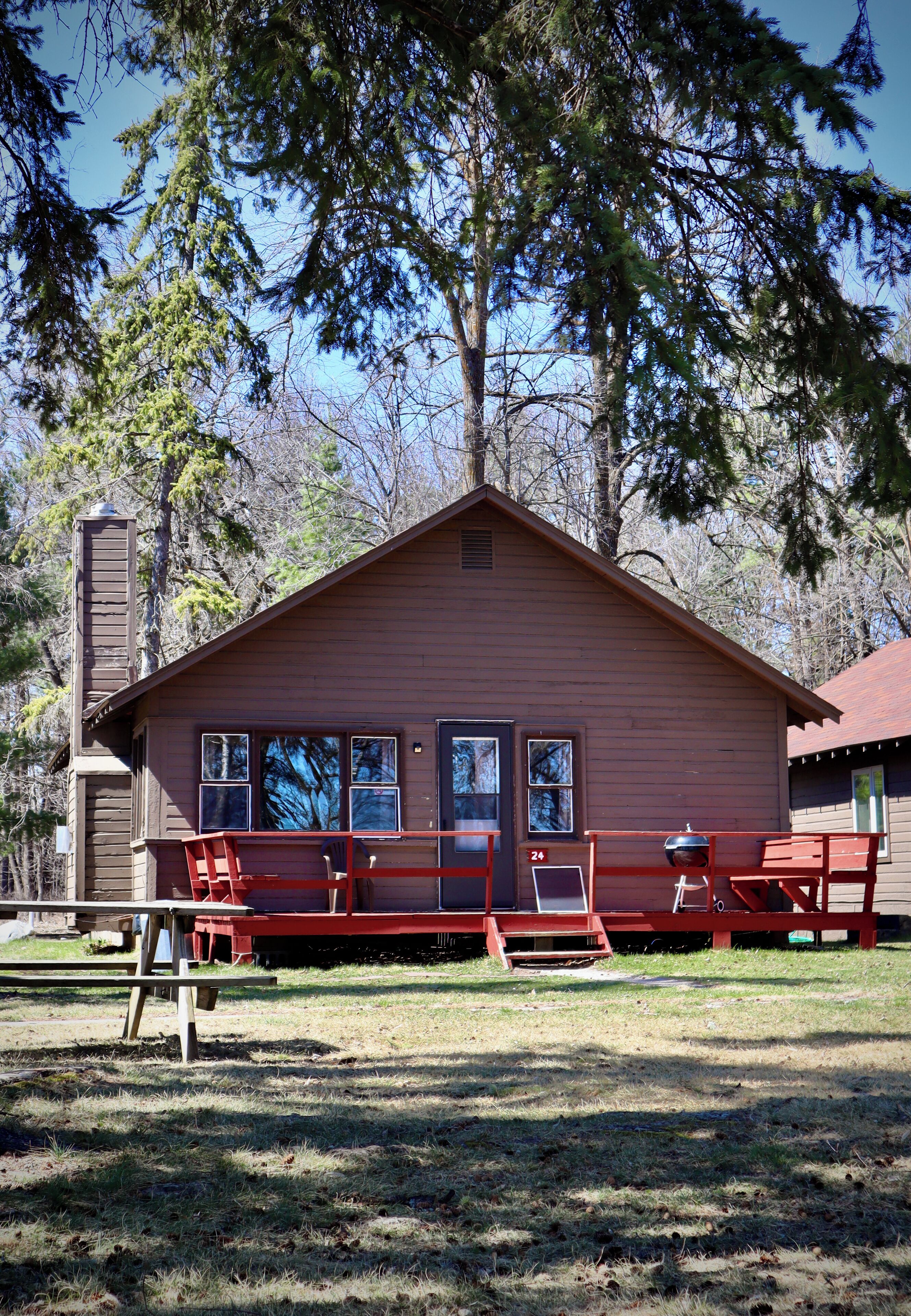 A rustic cabin with a chimney, a porch with red furniture, and a clear view of the surrounding trees.