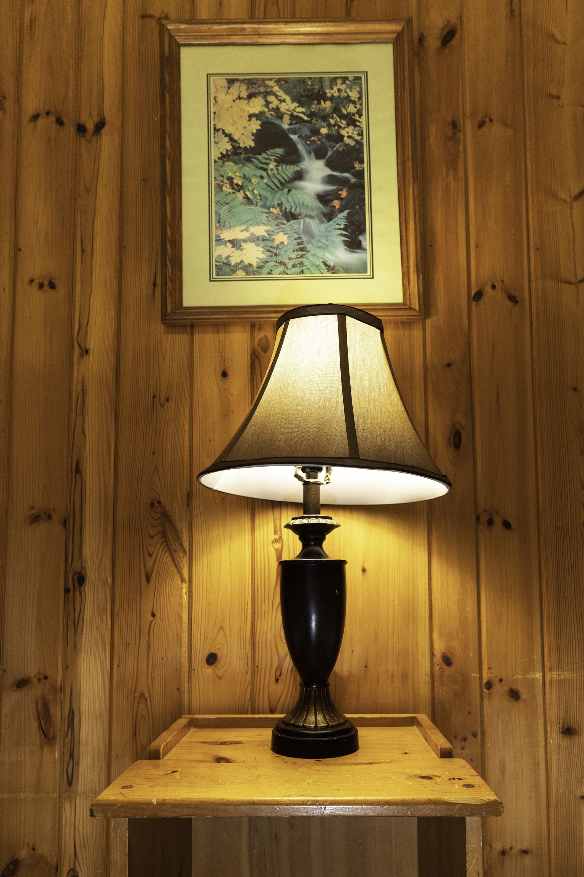 A table lamp with a black base and a white lampshade, placed on a wooden table against a wooden wall with a framed picture above it.