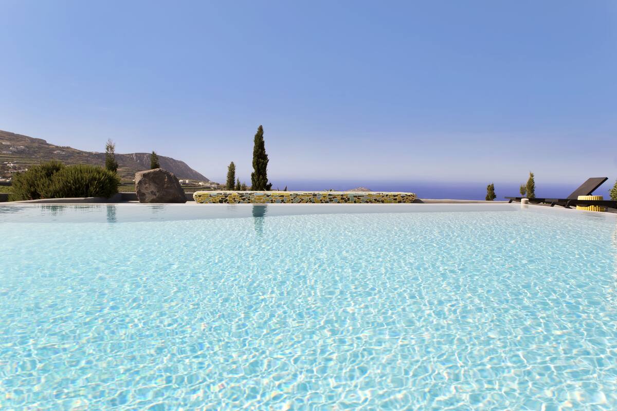 A clear blue pool with a stone border, surrounded by greenery and overlooking a mountainous landscape.