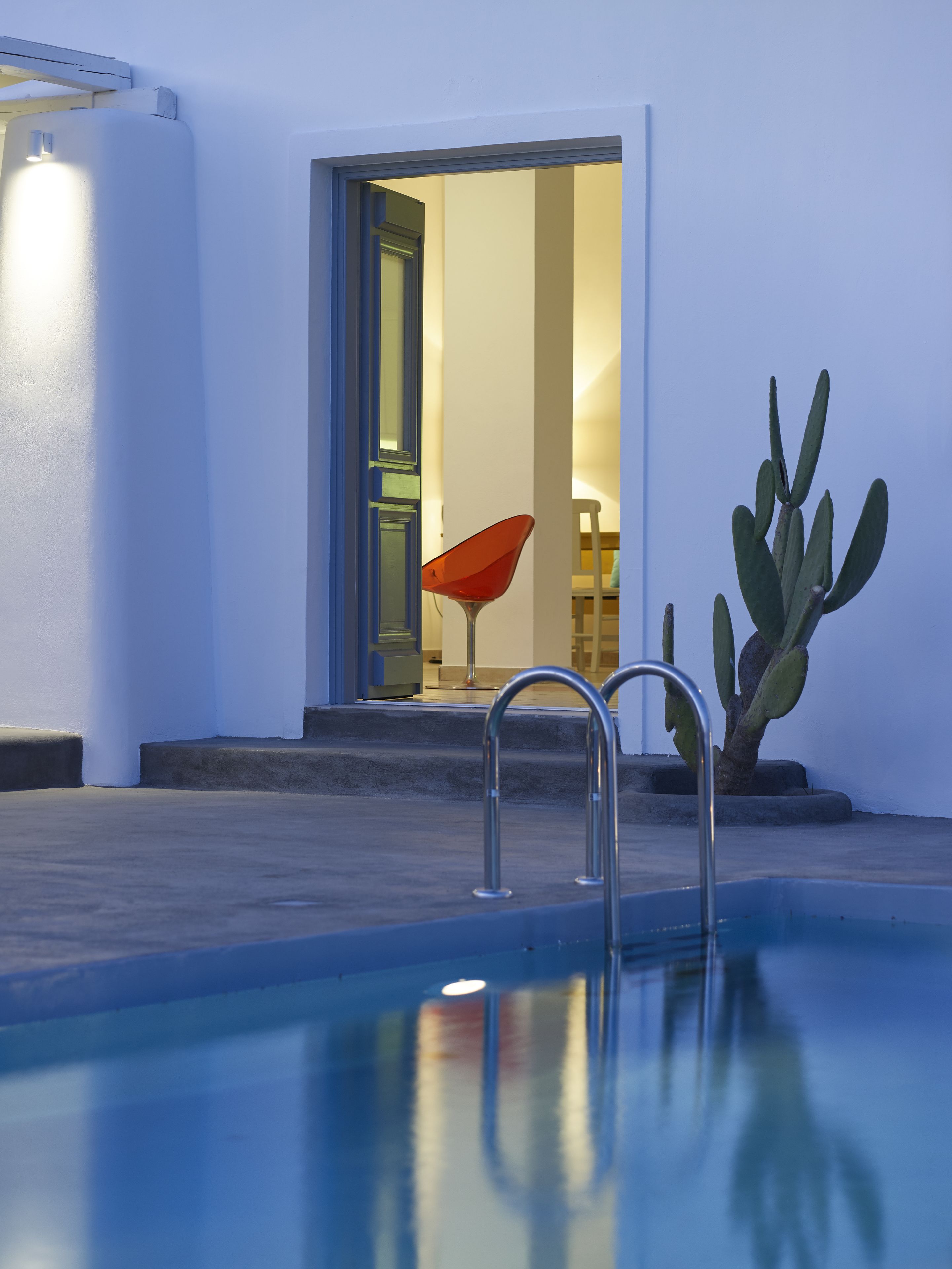 A swimming pool with metal ladders, a cactus plant, and a red chair near a doorway leading to an interior space.