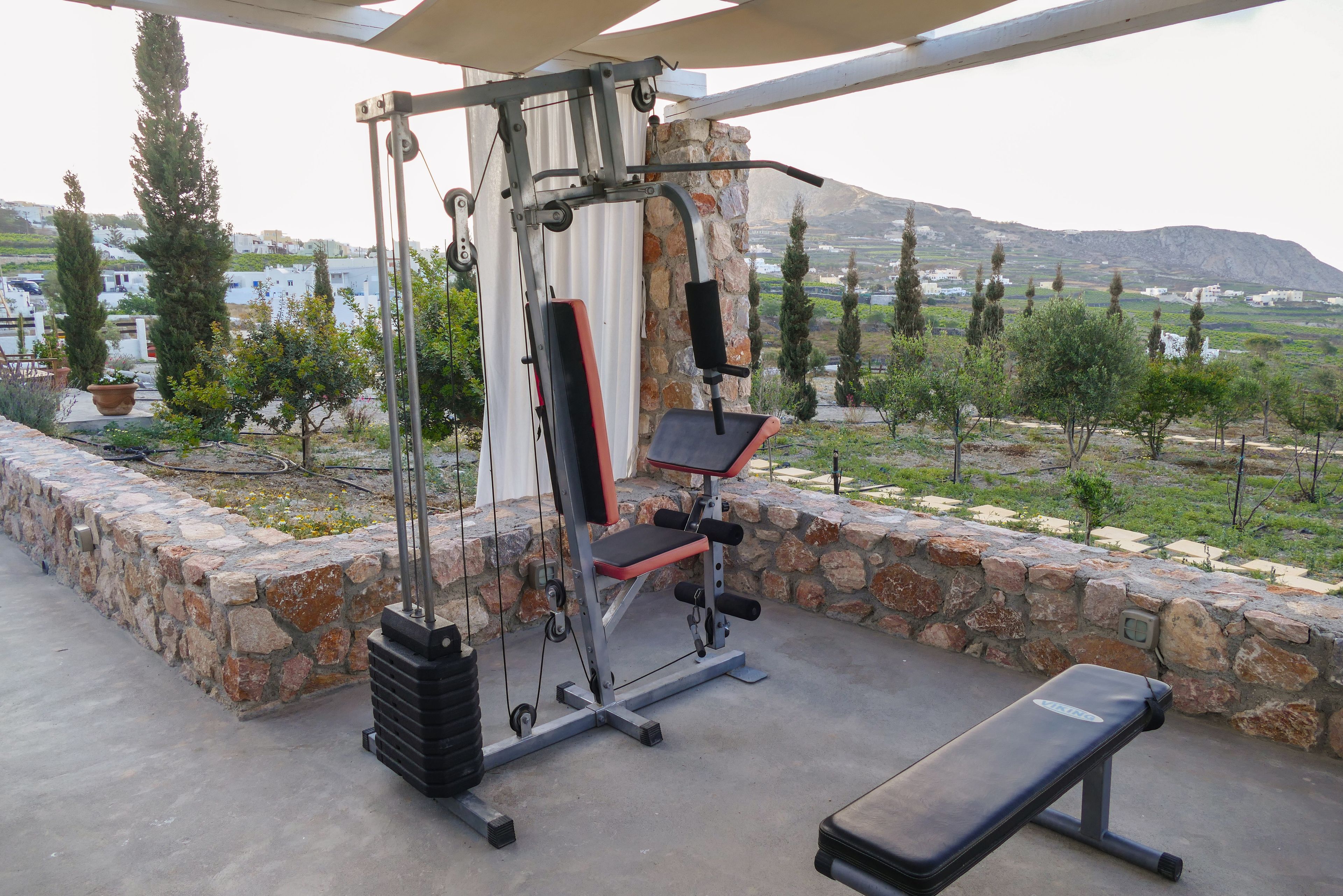 Un gimnasio al aire libre con banco y máquina de pesas, frente a una pared de piedra con vistas a un paisaje verde y edificios lejanos.