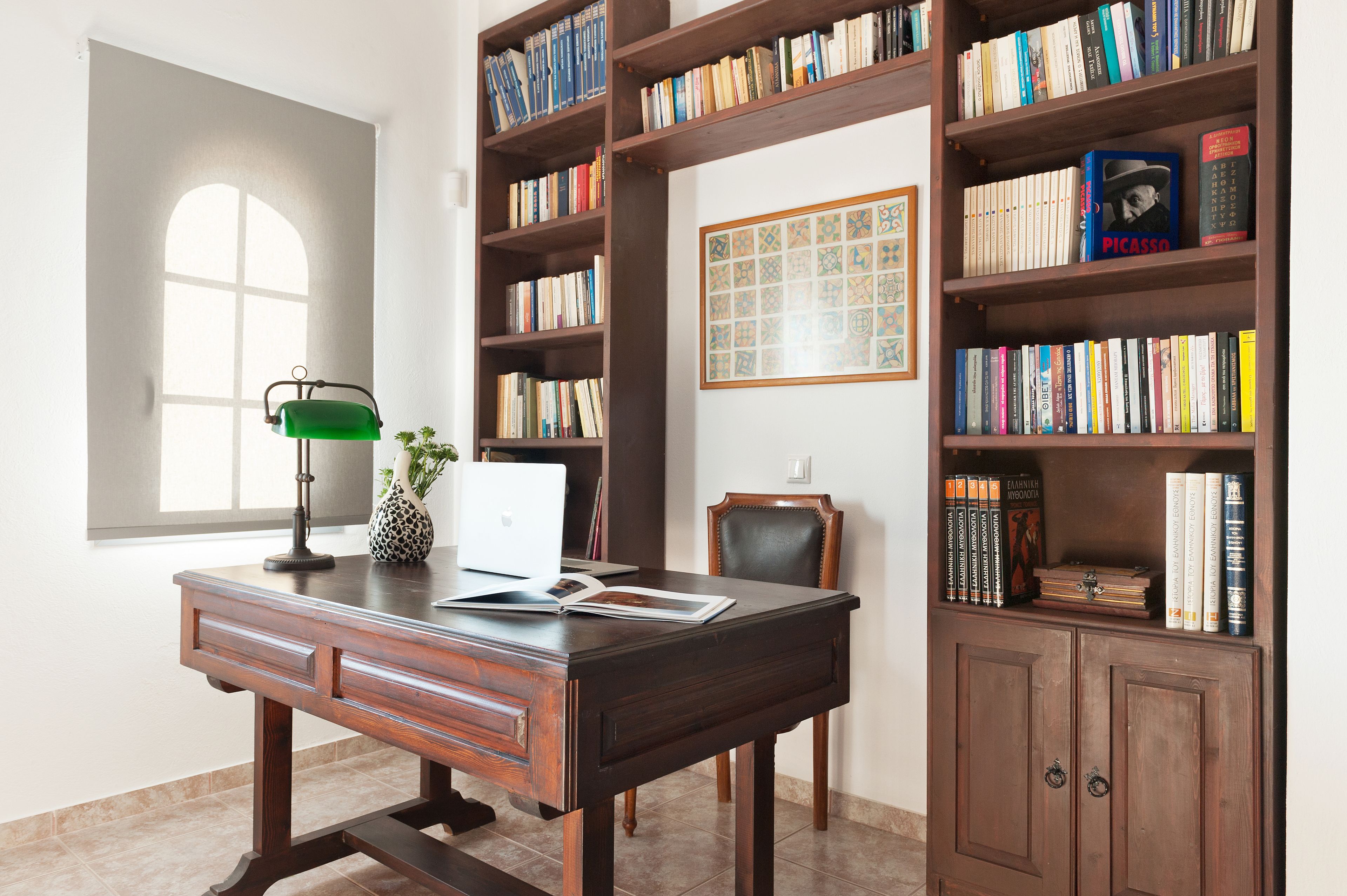 A study room with a wooden desk, bookshelves, a laptop, and a green desk lamp.