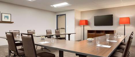 Un salón de conferencias con una larga mesa, sillas, un televisor de pantalla plana, dos lámparas y un cuadro en la pared.