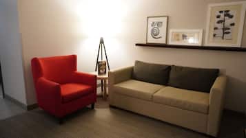 A room with a red chair, beige sofa, and a small table with a lamp. There are framed pictures on the wall.