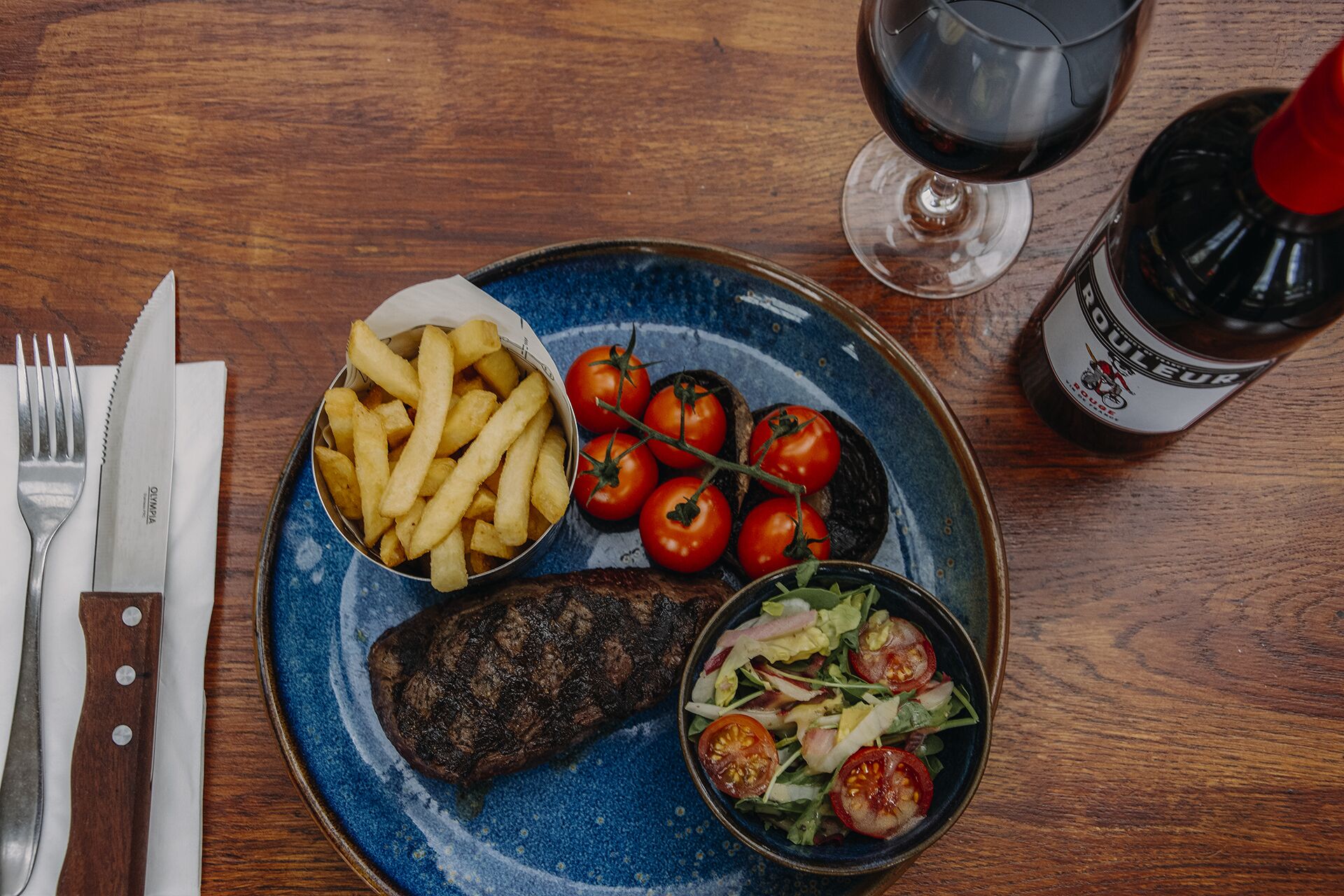 A plate with steak, fries, tomatoes, and salad, a glass of red wine, and a bottle of sauce on a wooden table.
