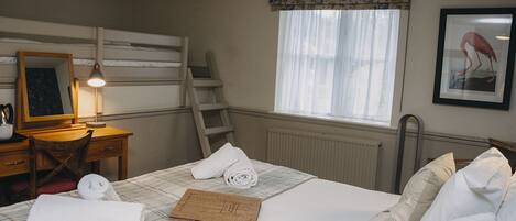 A hotel room with a bed, a desk with a lamp, a ladder, a window with sheer curtains, and a framed picture on the wall.