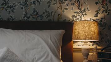 Un lit bien fait, avec une serviette enroulée, une lampe sur une table de chevet et un mur recouvert d’un papier peint à motifs floraux.