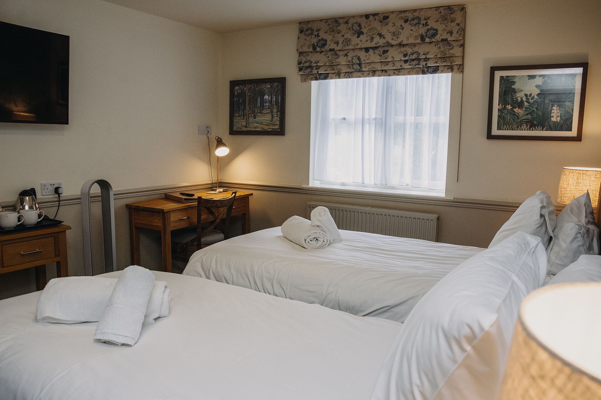 Une chambre d’hôtel avec deux lits, un bureau, une chaise, un radiateur et une fenêtre avec des rideaux.