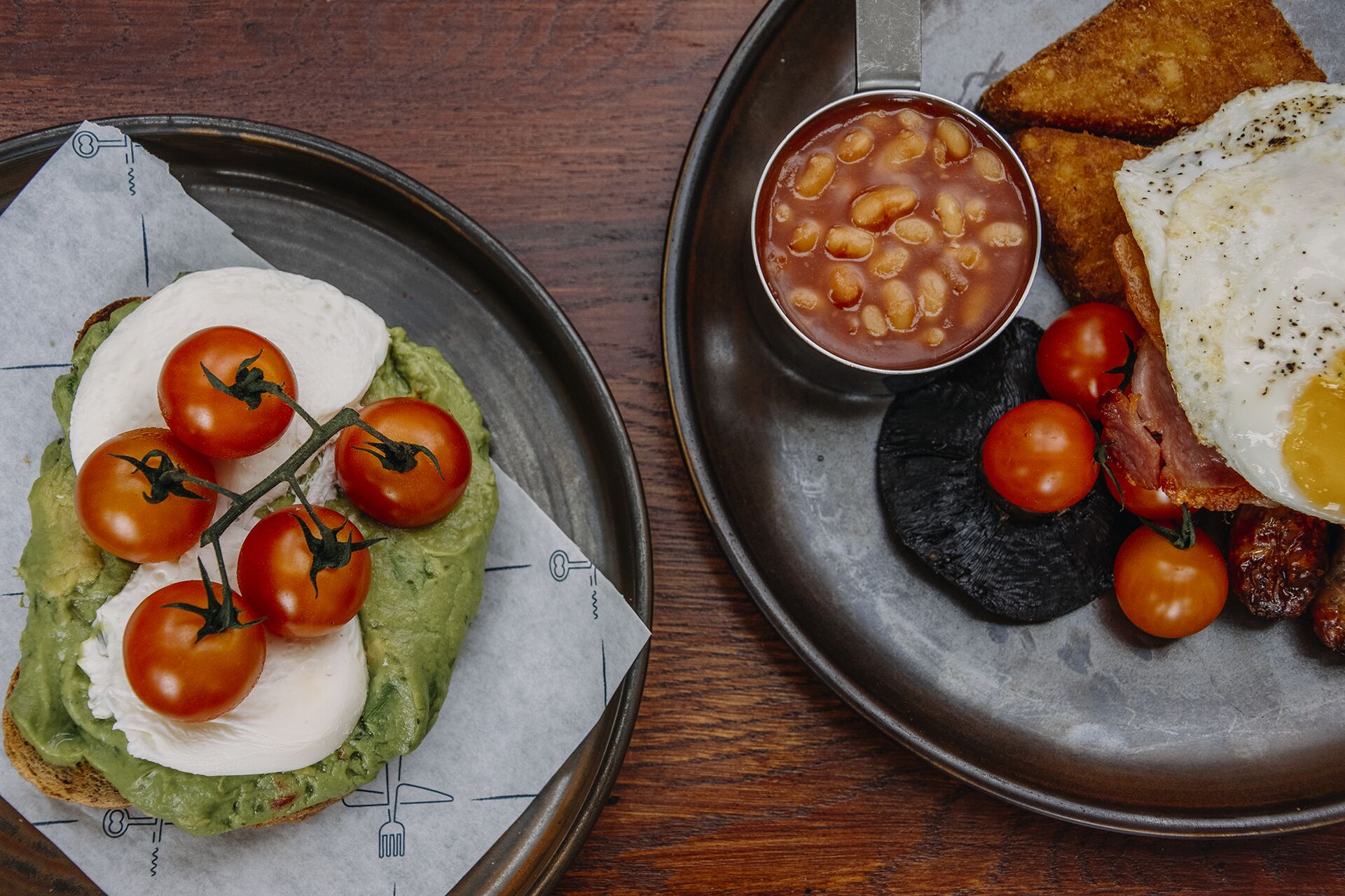 Un piatto per la colazione con pane tostato all'avocado, pomodori e formaggio, una ciotola di fagioli, un uovo fritto, pancetta e pomodorini.