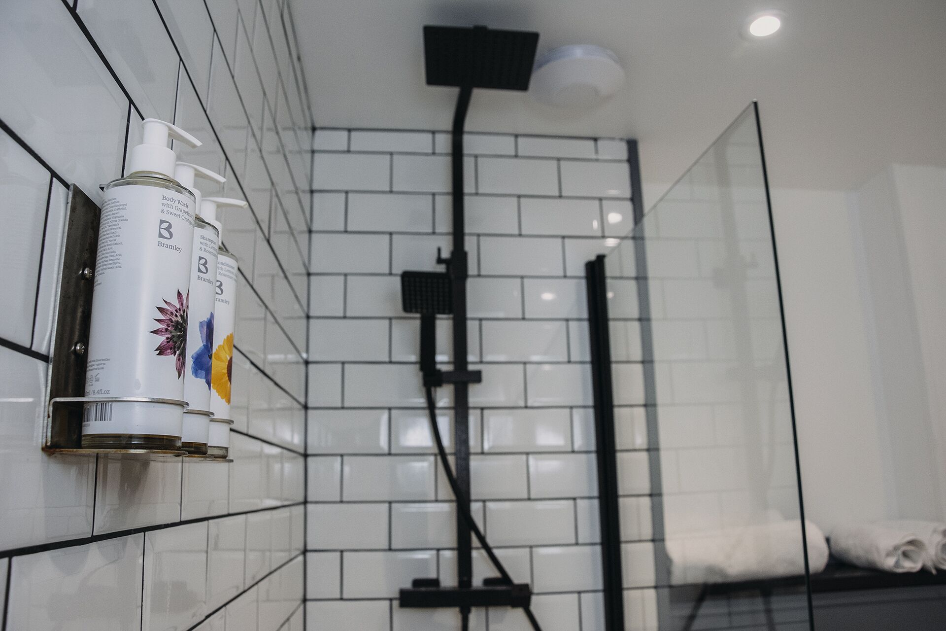 A bathroom with white tiled walls, a shower area, and a bottle of body wash on the wall.
