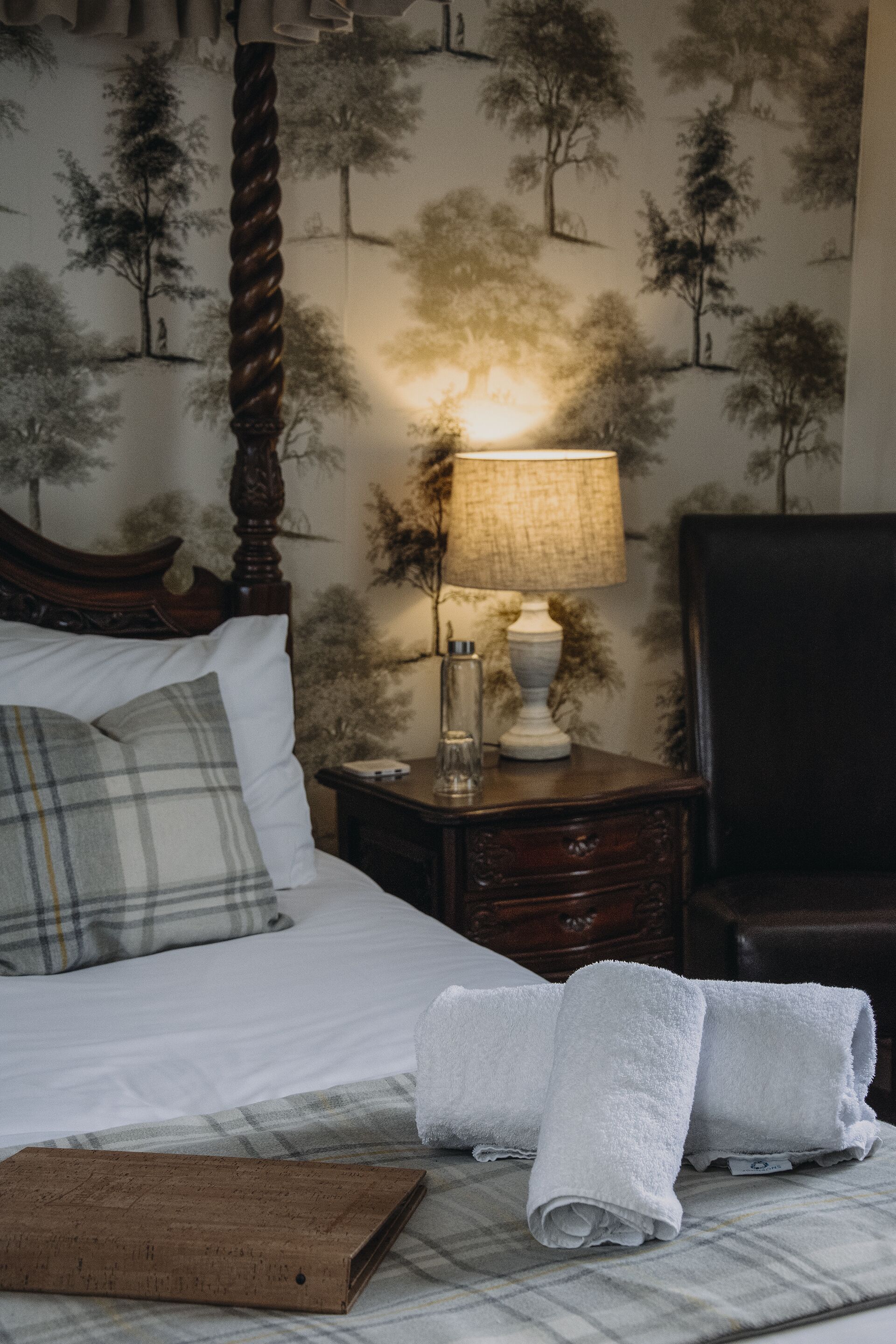Een net opgemaakt bed met witte lakens en een ruitkussen, een houten nachtkastje met een lamp, en een muur met een boompatroon.