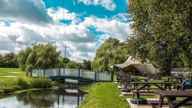 A riverside picnic area with a bridge, picnic tables, and a tent. - The George Inn (Warminster)