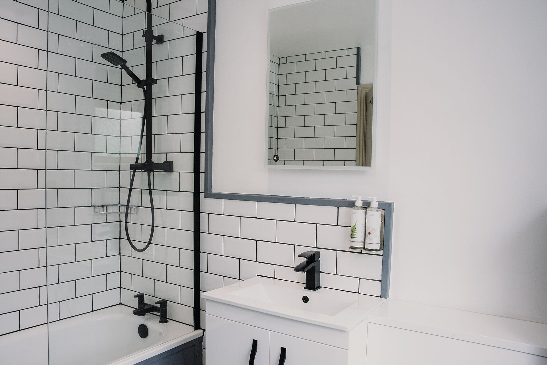 A modern bathroom with white subway tiles, a black showerhead, a white sink, and a mirror above the mirror cabinet.