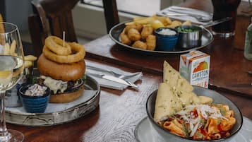 Una mesa de comedor con comida que incluye una hamburguesa, aros de cebolla y pasta. Hay salsas y una bebida sobre la mesa.