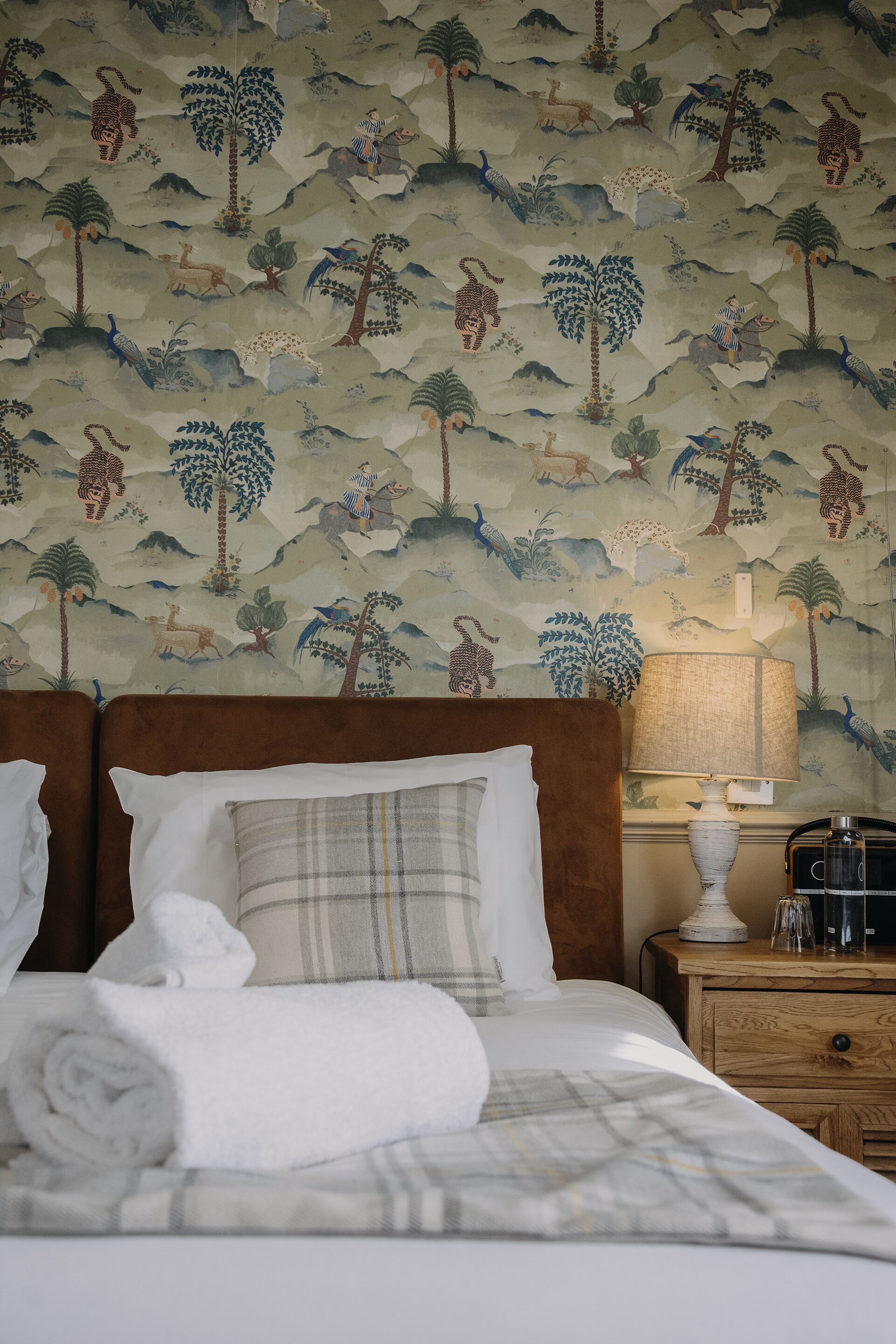 A hotel room with a bed, pillows, a nightstand, a lamp, and a wall with a jungle-themed mural.