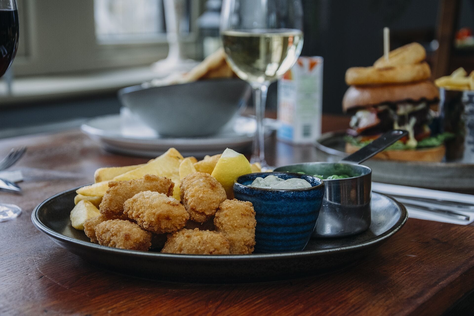 Een bord met gefrituurd en gepaneerd eten, een kom blauwe saus en een glas witte wijn op een houten tafel.