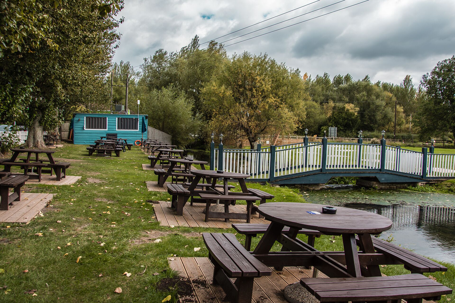A lakeside picnic area with wooden tables and benches, a small blue cabin, and a bridge over the water.