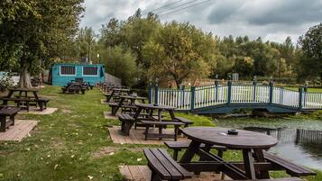 A lakeside picnic area with wooden tables and benches, a small blue cabin, and a bridge over the water.