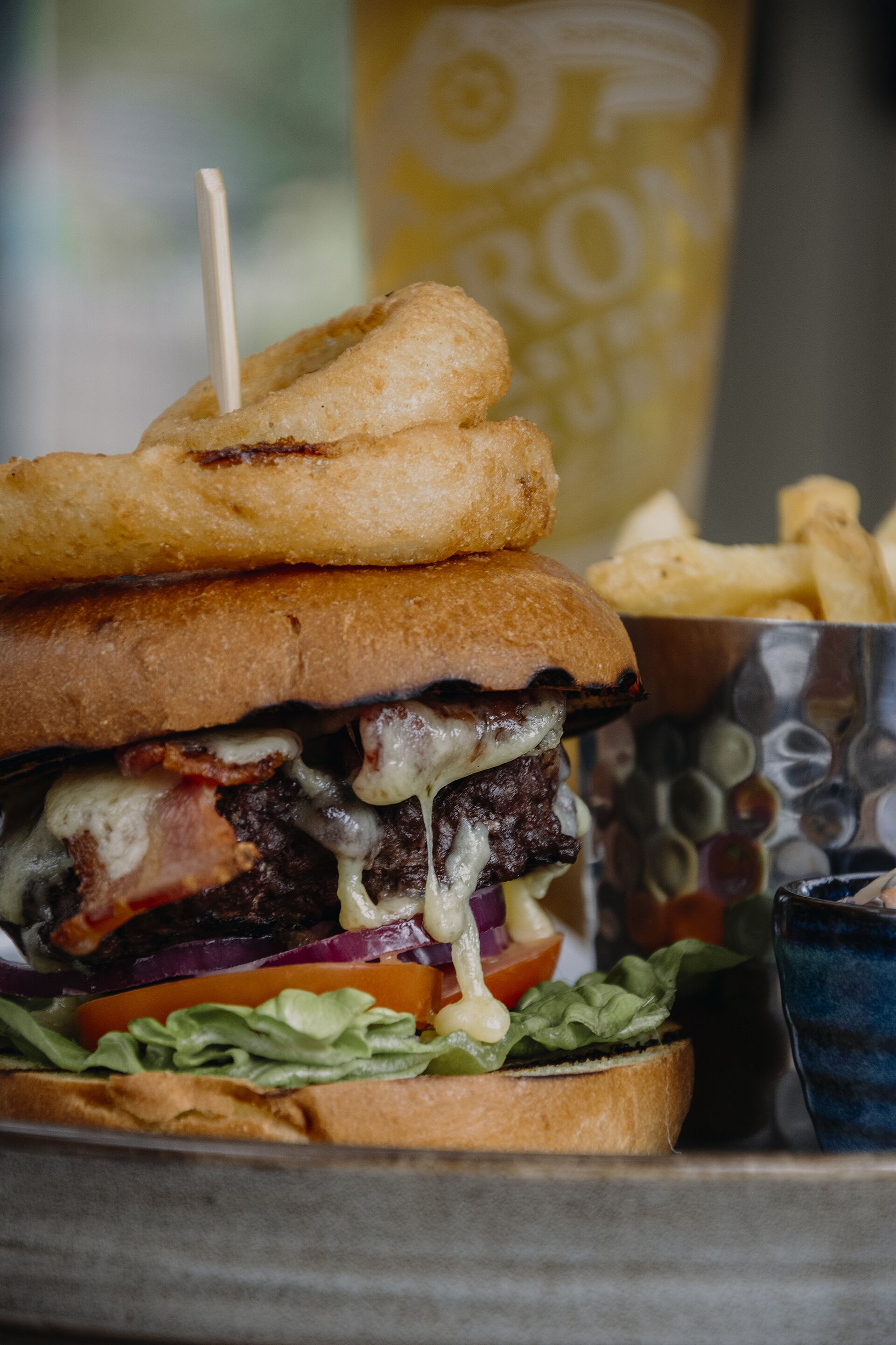 A burger with a toasted bun, beef patty, cheese, bacon, lettuce, tomato, and onion, accompanied by a side of fries and a drink in a branded glass.