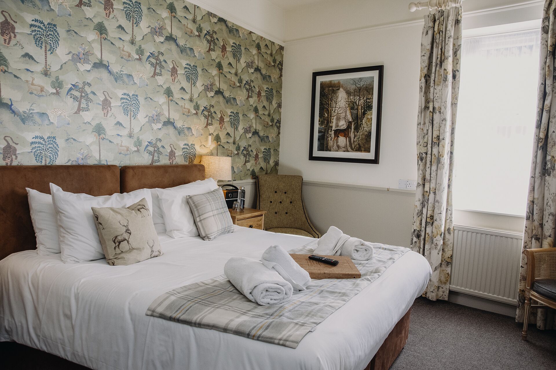 Een net opgemaakt bed met witte lakens en een geruit plaid, een houten nachtkastje met een lamp, een lijst met een foto aan de muur, en een raam met bloemen gordijnen.