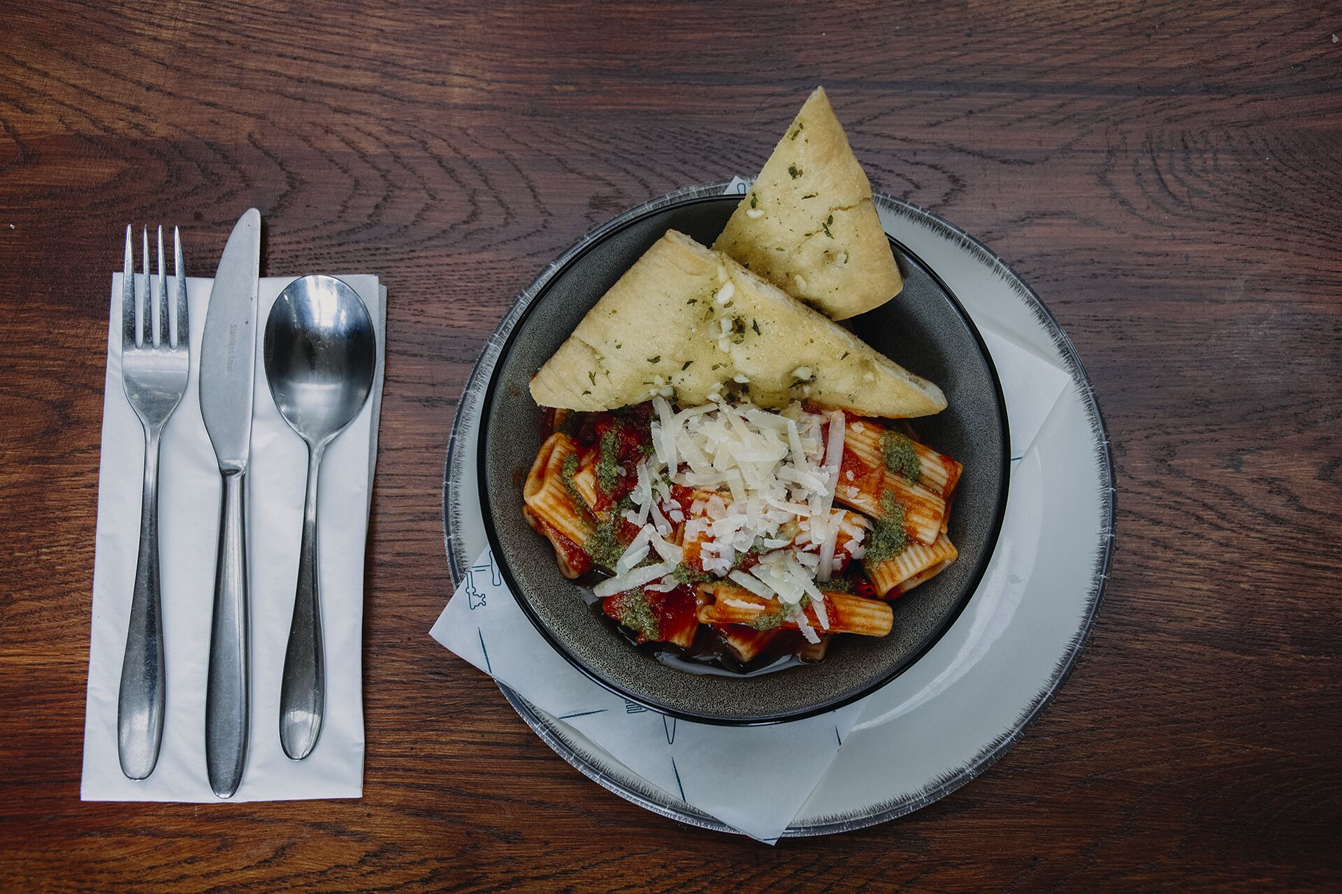 Un plato de pasta con queso y hierbas, acompañado de un tenedor, cuchillo y cuchara sobre una mesa de madera.
