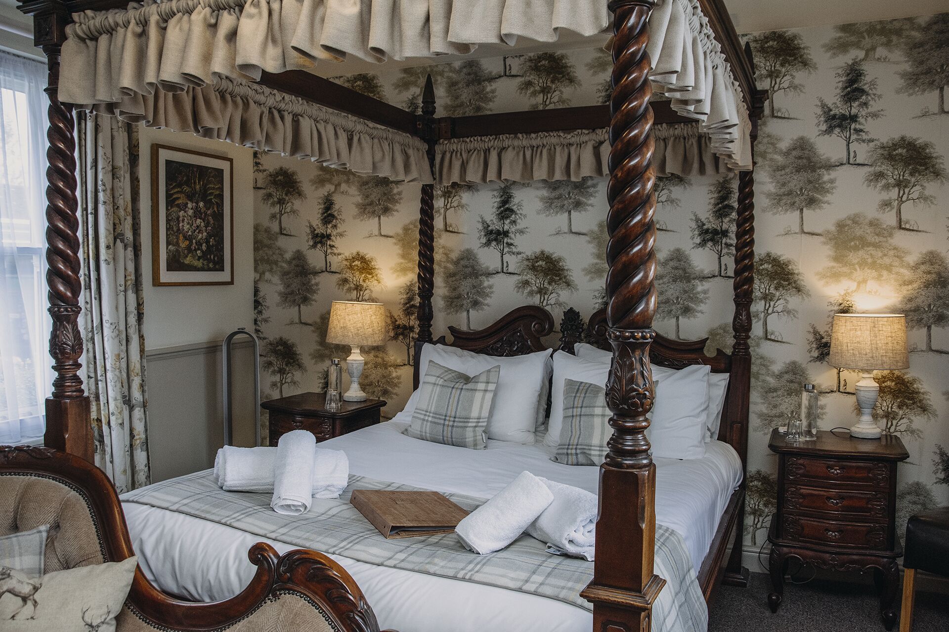 A four-poster canopy bed with white linens, two bedside tables with lamps, and a wall with a tree pattern.