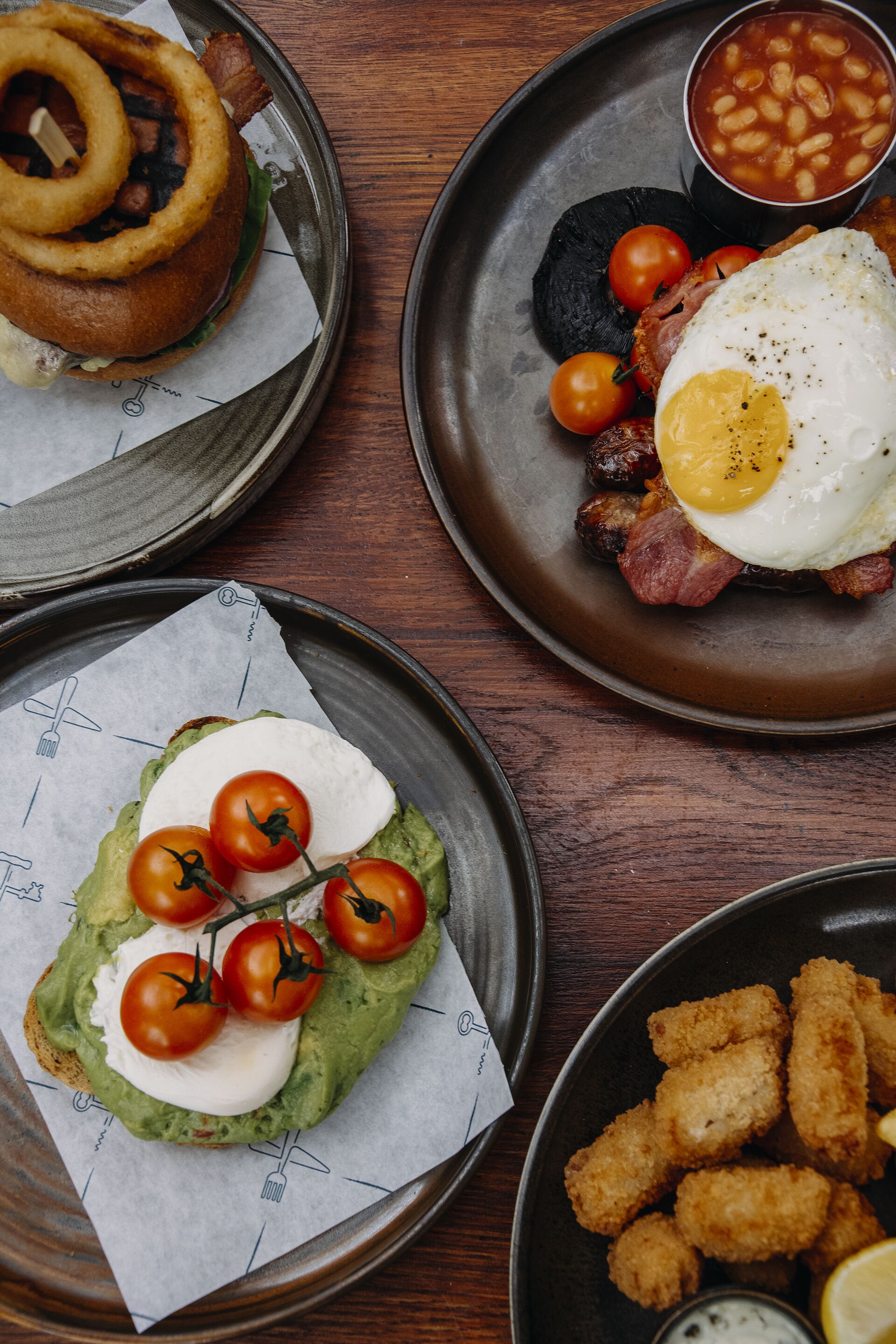 Variedad de platos que incluyen una hamburguesa, huevos, frijoles, tostada de aguacate y opciones fritas, sobre una mesa de madera.