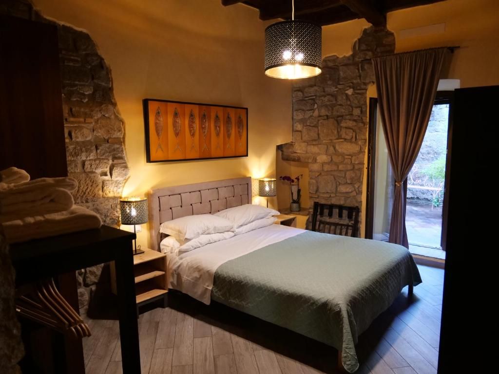 A bedroom with a stone wall, a bed with a headboard, a nightstand, a lamp, and a window with curtains.