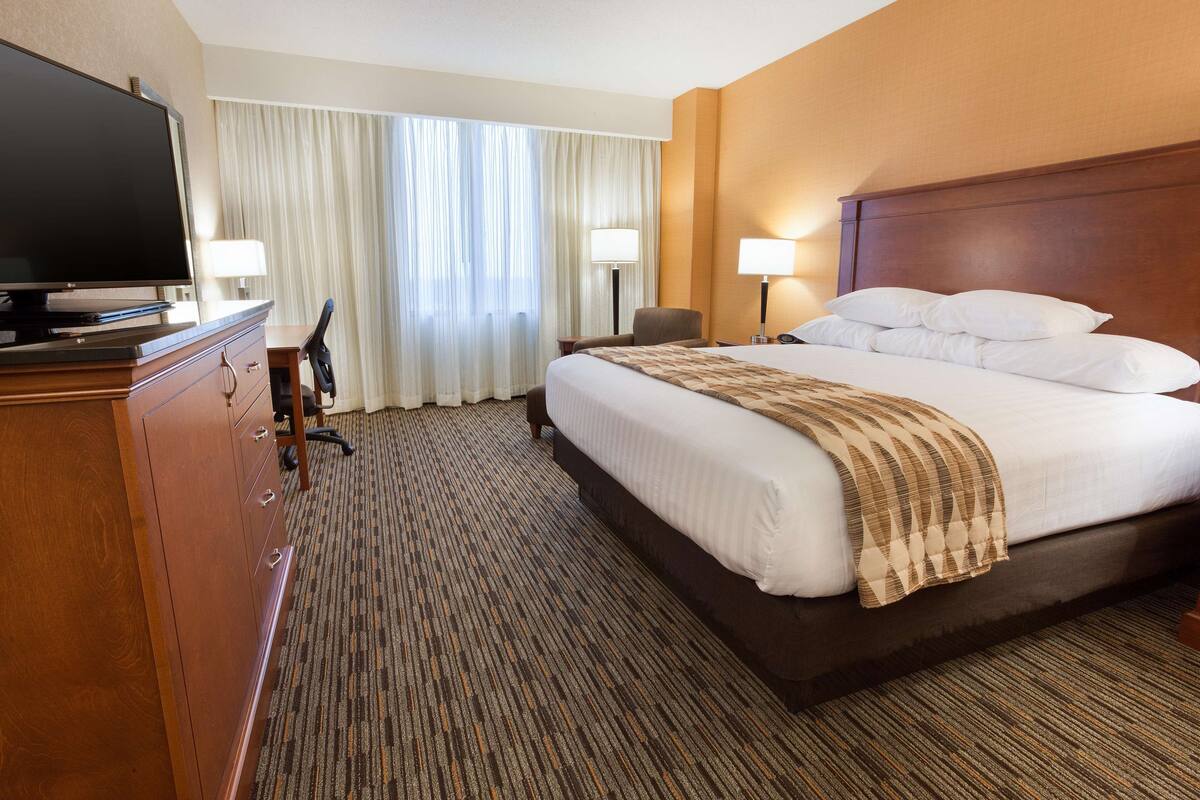 A hotel room with a large bed, a television on a wooden dresser, a desk with a chair, and two lamps.