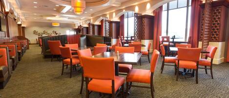 Un comedor de hotel con varias mesas y sillas color naranja, suelo alfombrado e iluminación decorativa en el techo.