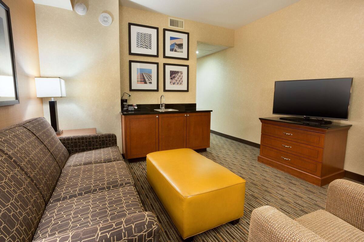 A hotel room with a sofa, ottoman, television, and a kitchenette area.