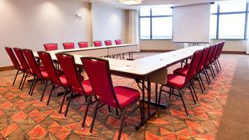 Un salón de conferencias con una larga mesa y sillas rojas, ventanas grandes y una pantalla de proyector.