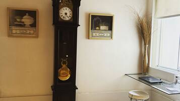 A room with a tall grandfather clock, a small stool, a glass table, and two framed pictures on the wall.