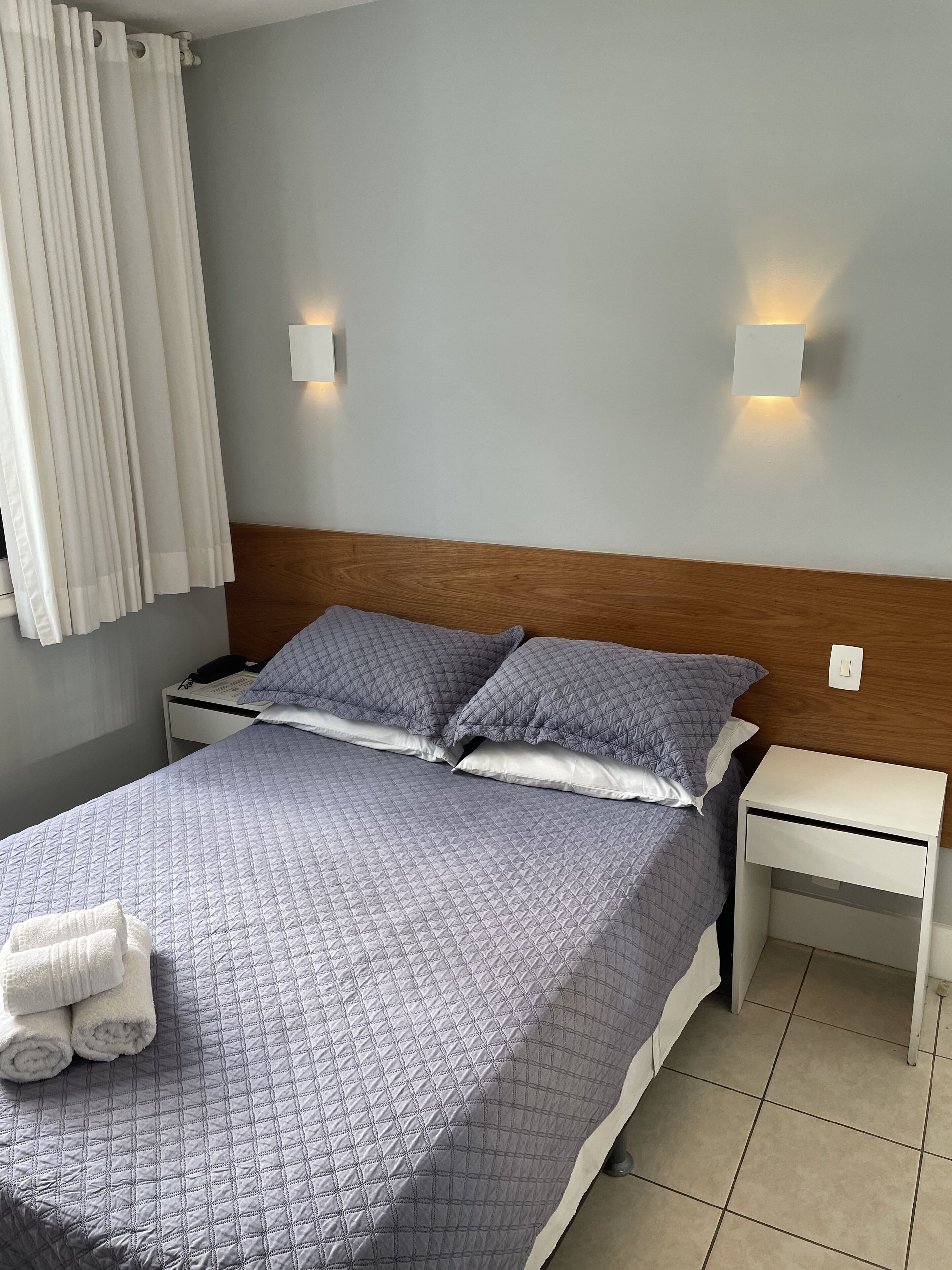 Een netjes opgemaakt bed met een gewatteerde hoes, twee kussens, een nachtkastje met een lamp en een wandlamp.