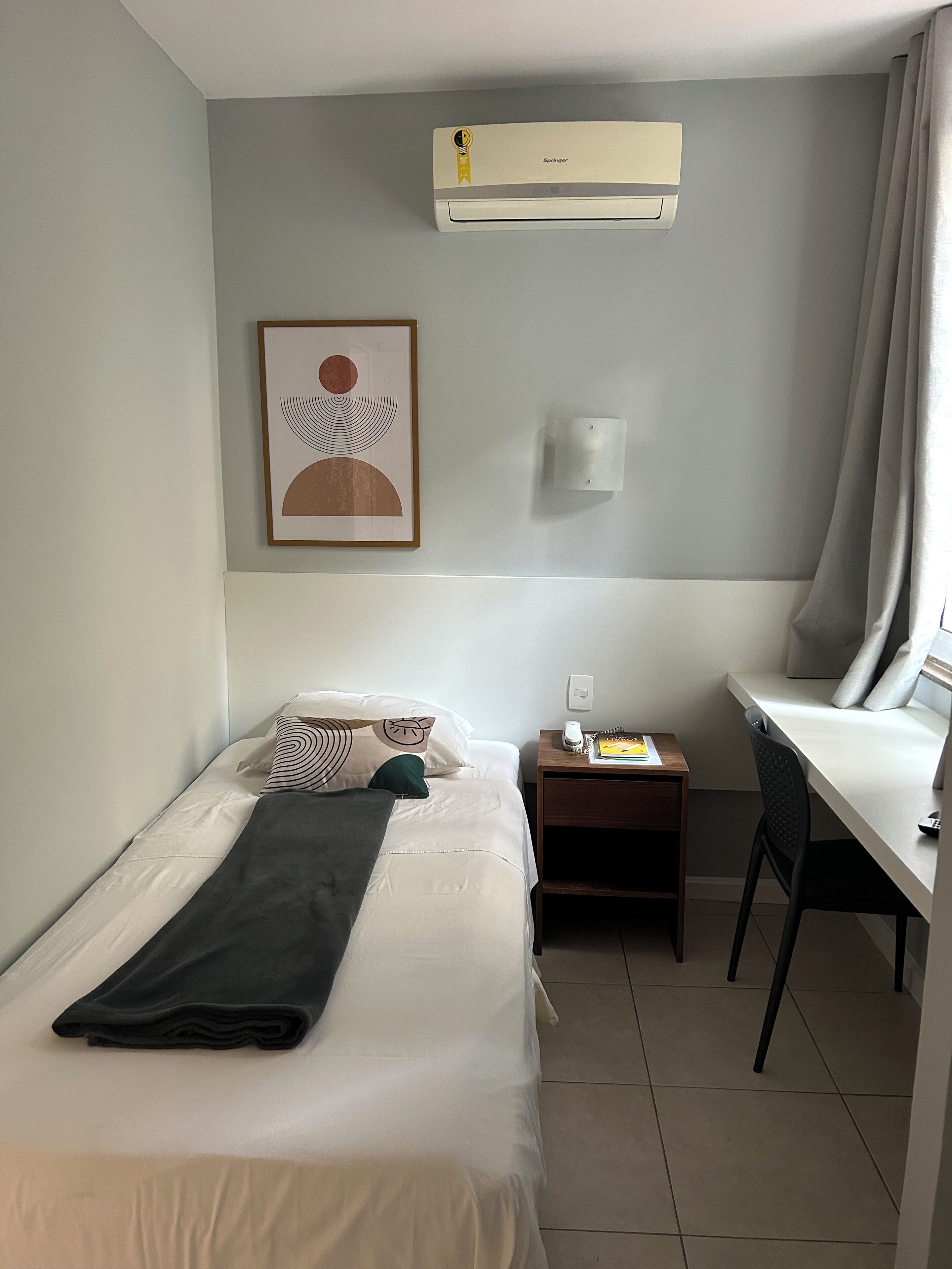 Hotelkamer met een bed, een bureau, een stoel, een airconditioner, een wandlamp en een kunstwerk in een lijst.