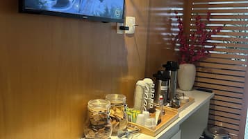 Een moderne hotel receptie met een flatscreen televisie aan de muur, een koffiehoek met diverse dranken en snacks, en een prullenbak.